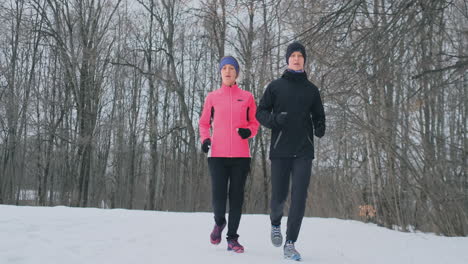 Young-man-and-woman-on-a-morning-run-in-the-winter-forest.-A-woman-in-a-loose-jacket-a-man-in-a-black-jacket-is-running-through-a-winter-park.-Healthy-lifestyle-happy-family.