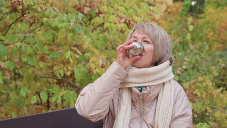 middle aged lady in beige winter coat and knitted cream scarf sits on wooden park bench during autumn afternoon, sipping hot coffee from stainless cup while daughter and dog relax among golden leaves