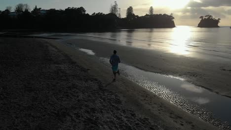 aerial follow shot of silhouetted young man running on a beach in auckland, new zealand