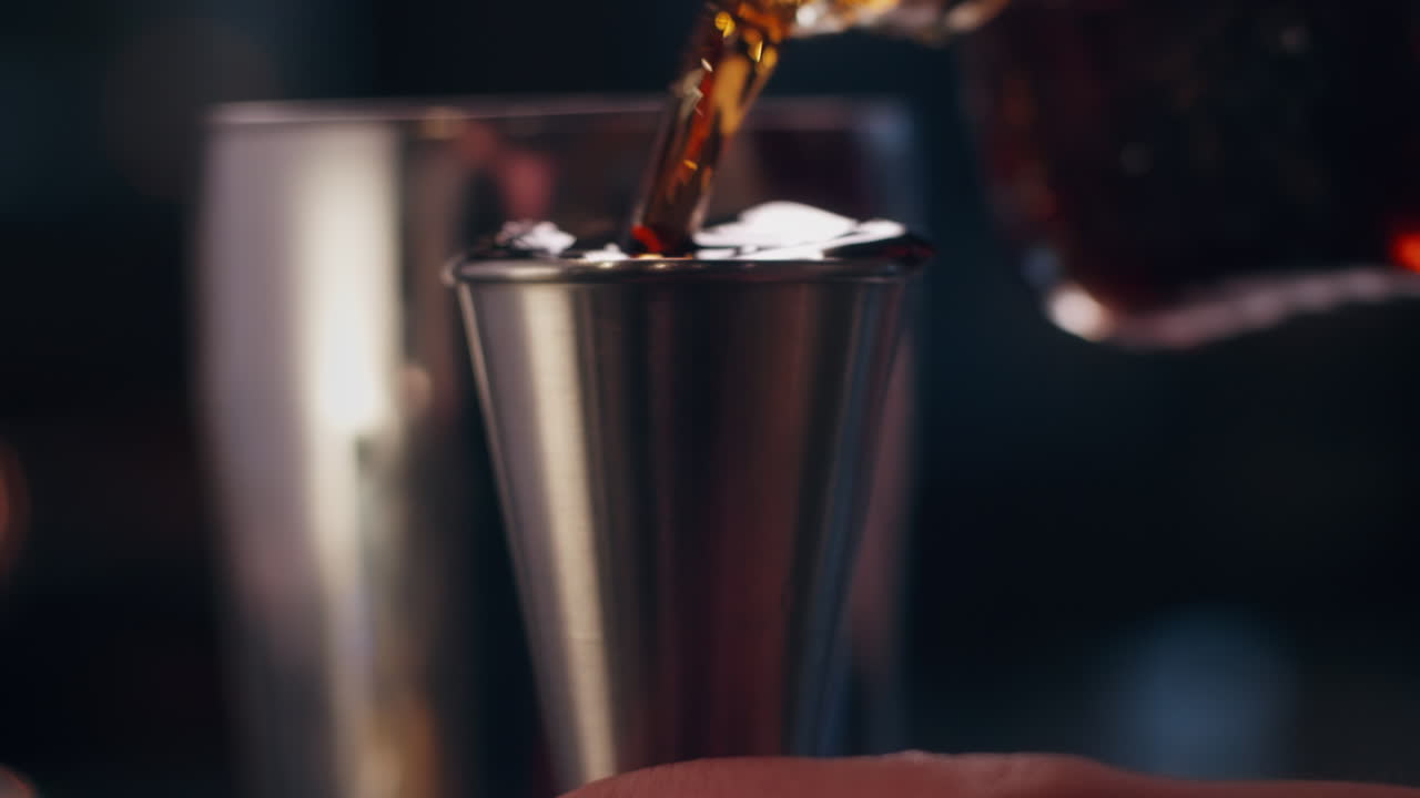 Primo piano di un distillato scuro versato in un misurino di metallo in un cocktail bar con luci soffuse.