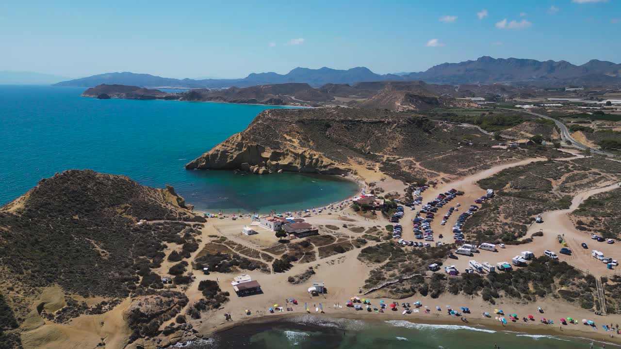 Aerial view of Playa de la Carolina on the Águilas coastline in the Murcia region of Spain, showing the sandy cove, clear water, rugged cliffs, parked cars and beach visitors on a sunny day