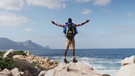 rear view of african american man standing on rock with arms wide open while trekking in the mountai