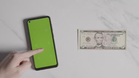 Overhead-Currency-Shot-Of-US-5-Dollar-Bill-Next-To-Person-Using-Green-Screen-Mobile-Phone