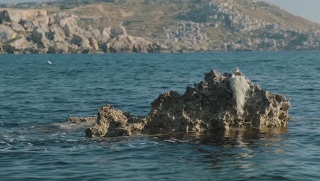 water gently splashes on a rock formation in the middle of the sea with an arid, stony mountain landscape in the background