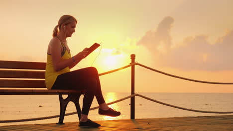 Fitness-Mujer-Disfruta-De-Una-Tableta-Se-Sienta-En-Un-Banco-En-Un-Muelle-Contra-El-Telón-De-Fondo-Del-Sol-Naciente-4k-Vi