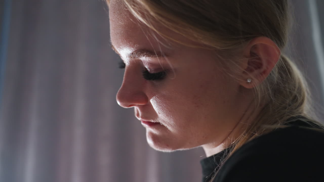 Close up of female massage therapist with tied hair and focused expression, concentrating intently during therapy session with soft light highlighting her facial features against gray curtain