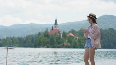 young woman using phone by the lake with mountains and church in background