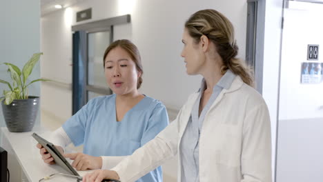 focused diverse female doctors using tablet and discussing at hospital reception desk, slow motion