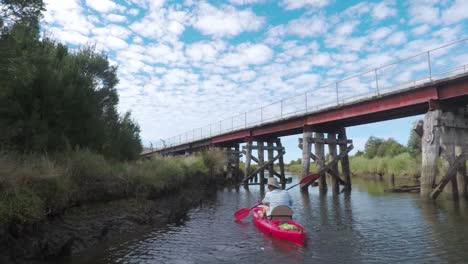 Tracking-shot-of-a-man-in-a-red-kayak-paddling-down-a-river-with-an-old-bridge-extending-over-it