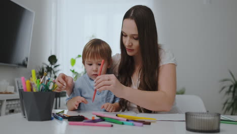 Sentada-En-La-Mesa-Del-Salón,-Una-Joven-Madre-Le-Enseña-A-Su-Hijo-A-Sostener-Un-Lápiz-Y-Le-Enseña-A-Dibujar