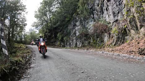 group of motorcyclists riding on a gravel road surrounded by forests and rock walls. chilean patagonia