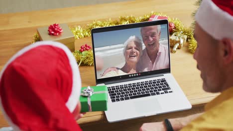 Kaukasischer-Vater-Und-Sohn-Mit-Weihnachtsmützen-Nutzen-Laptop-Für-Weihnachtsvideoanruf-Mit-Paar-Auf-Dem-Bildschirm