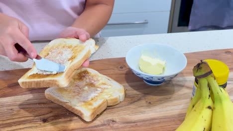 person spreads butter and dark spread on toasted bread slices at a kitchen counter, using a knife, under bright indoor lighting with a static camera