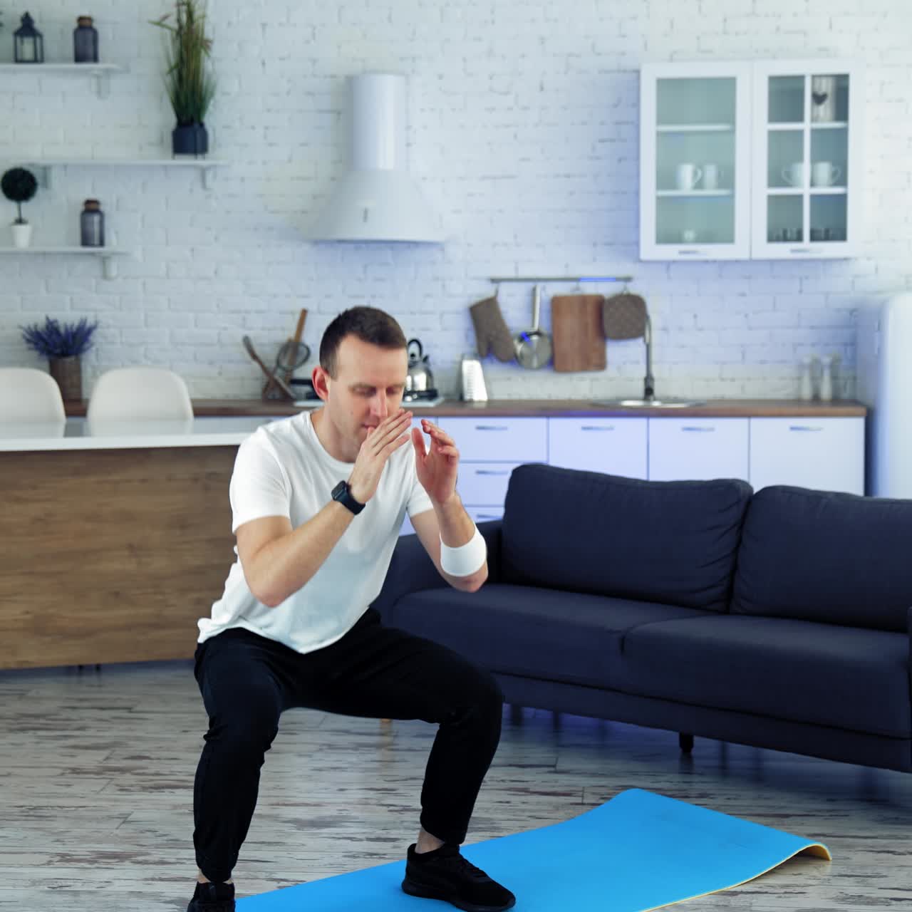 Home workout. Young man in white t-shirt and black pants doing squats exercises in kitchen interior. Sports training indoors. Slow motion