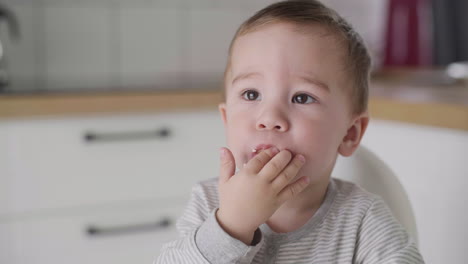 portrait of cute baby boy eating banana sitting in high chair in the kitchen