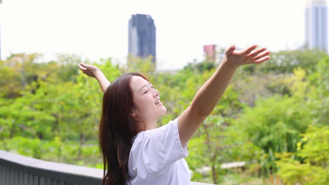 หญิงเอเชียสวมเสื้อยืดสีขาว ชูแขนขึ้นด้วยความดีใจ โดยมีฉากหลังเป็นสวนสีเขียวชอุ่ม