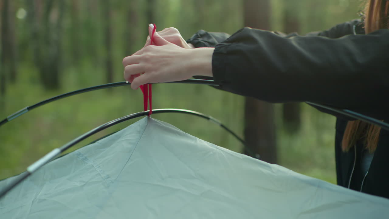 close up of woman hand tying red rope of tent to flexible black pole in forest environment while setting up shelter for camping with soft daylight and greenery in blurred background