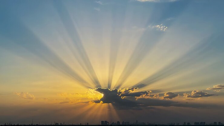 Sun sinking behind central dark cloud producing sunbeams across western horizon, city skyline