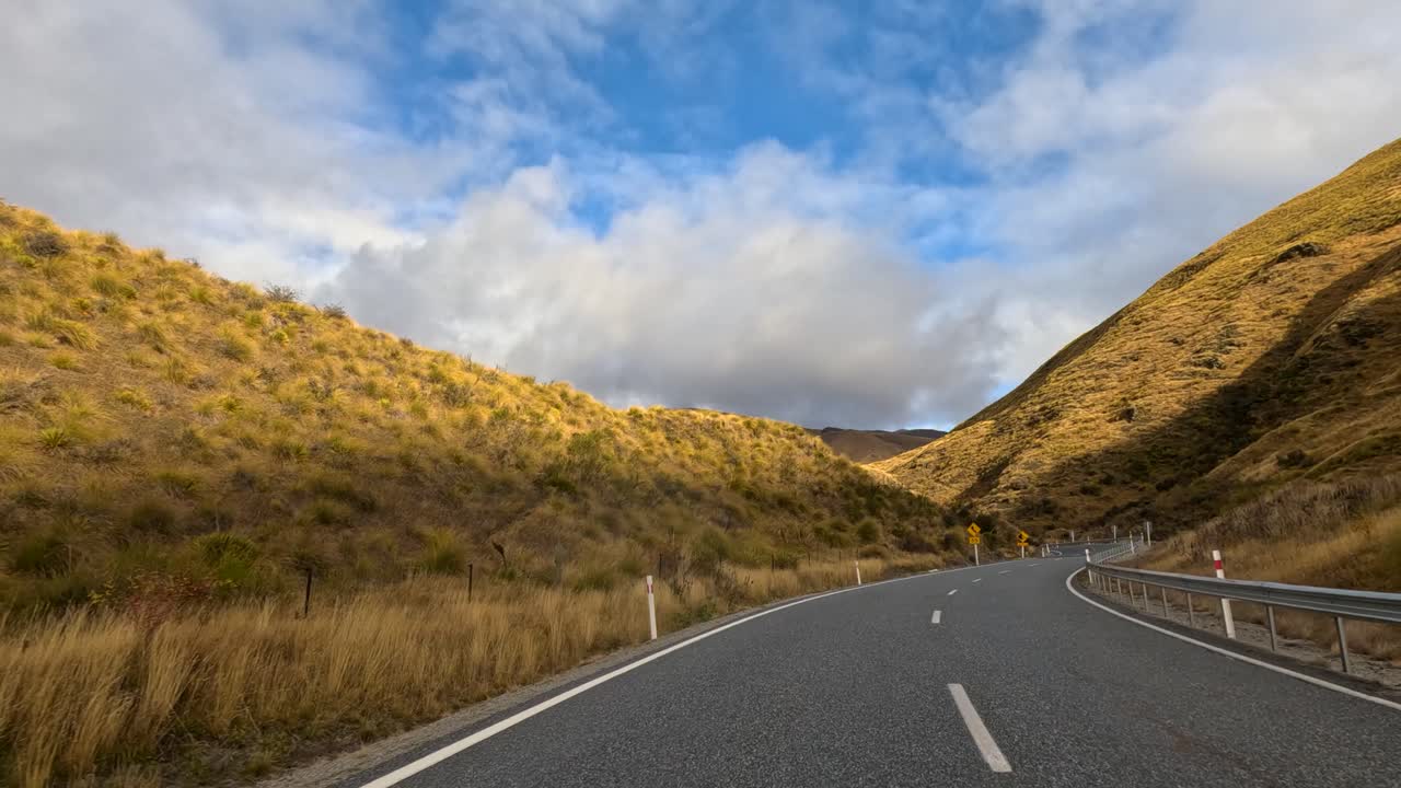 Point-of-view drive through sunlit rural hills on a paved mountain road, with road signs, guardrails, and dramatic clouds, captured in smooth daylight motion