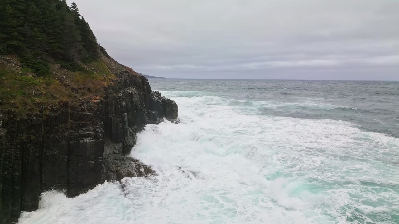 A drone captures Middle Cove’s massive sea cliffs as turbulent waves crash against the rocks, sending spray into the air under a grey, stormy sky