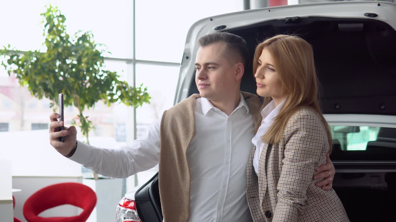 Happy couple taking a selfie at a car dealership with a new car in the background
