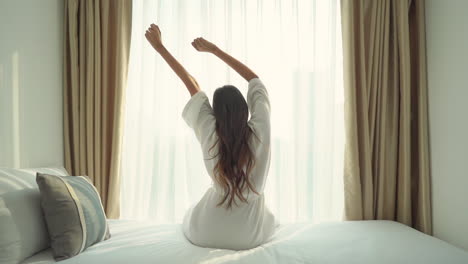 Beautiful-brunette-woman-sitting-on-bed-stretching-arms-on-bathrobe,-bright-window-background