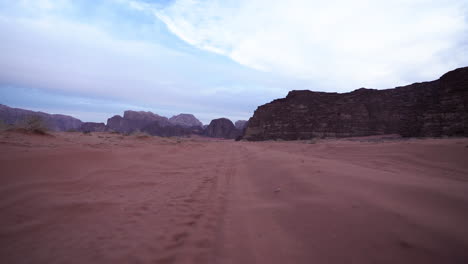 Toma-En-ángulo-Bajo-De-La-Cámara-Moviéndose-Por-El-Camino-De-Arena-En-El-Desierto-De-Wadi-Rum-Con-Nubes-Y-Cielo-Azul-En-El-Fondo