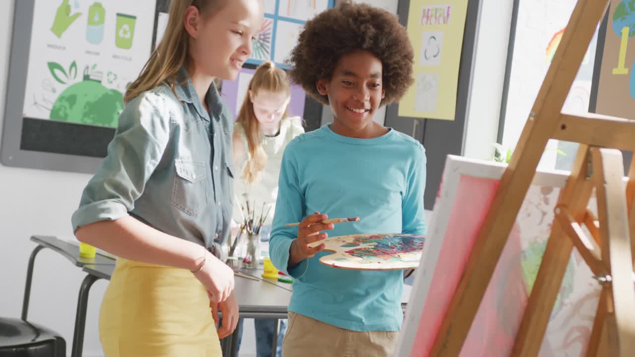 Video of happy diverse girl and boy talking by boy's easel in art class, copy space