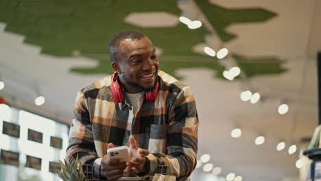 A-happy-man-with-Black-skin-in-a-checkered-shirt-and-red-wireless-headphones-leaning-on-a-cart-drives-through-the-supermarket-and-examines-the-goods-on-the-counter-and-selects-from-them-those-that-he-needs