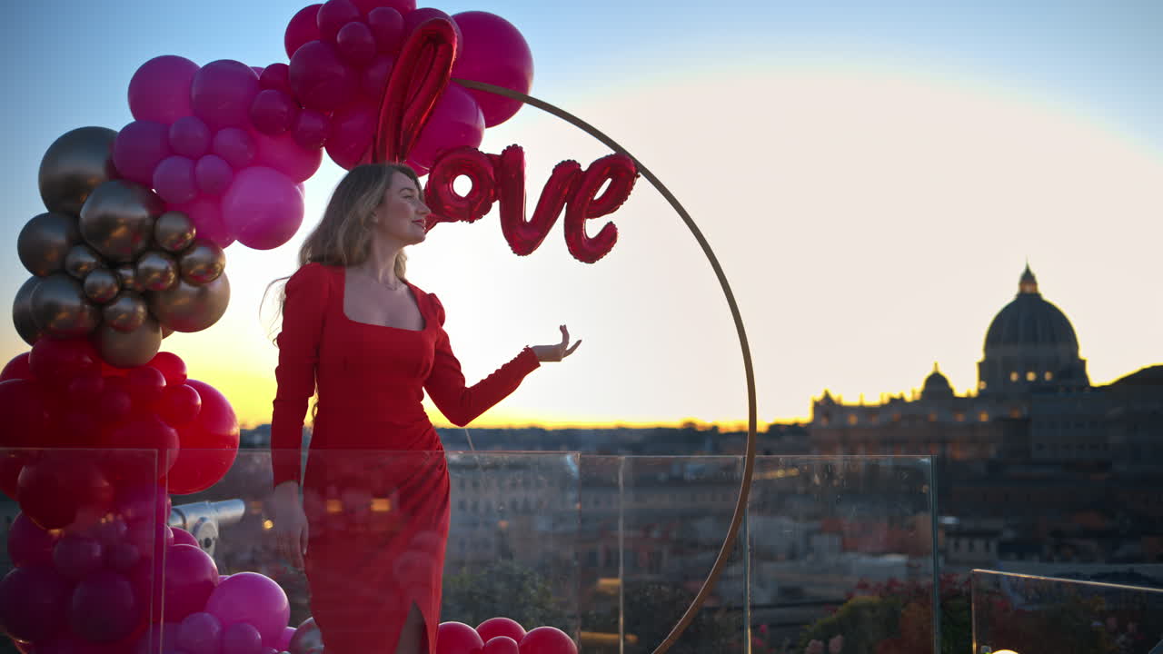 Beautiful young woman in red dress dancing at sunset near the red and pink balloon arch spelling out the word "love". Vatican city in background. Rome, Italy