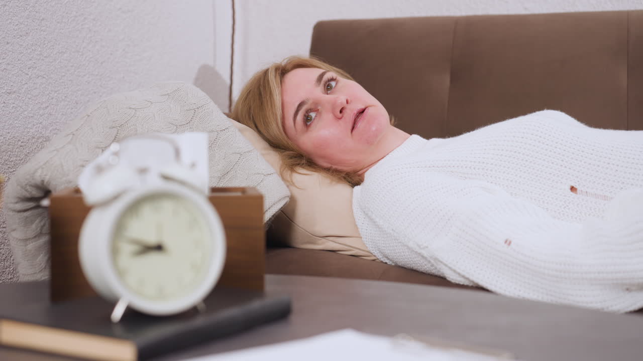Patient lies sideways on sofa facing counselor, conversing quietly, pillow supports head, table beside holds clock, tissue box, pencils, warm therapy room atmosphere, soft lighting