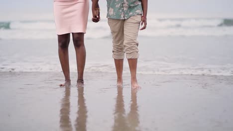 african american couple holding hands and walking in the beach, front view of two lovers with the sea behind