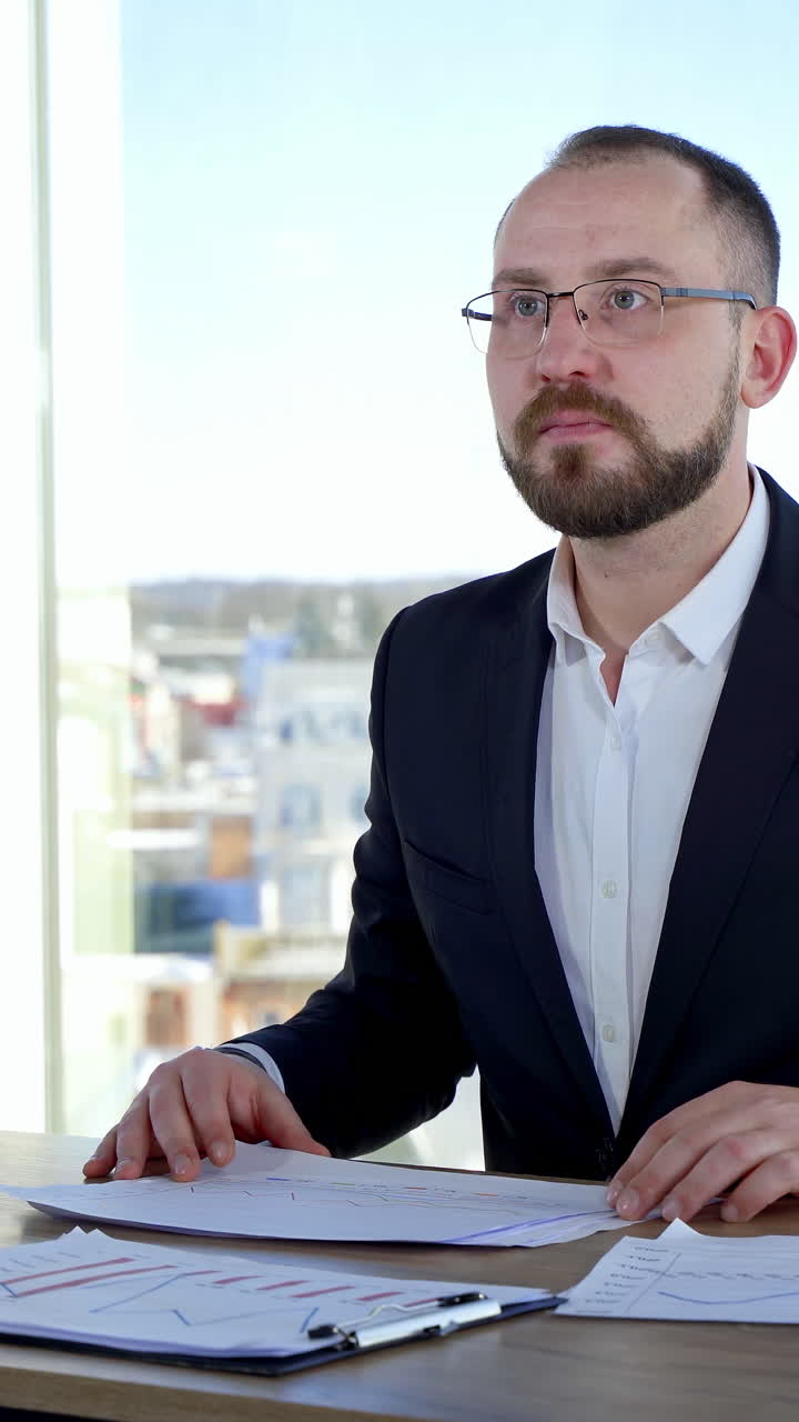 Businessman in glasses at his workplace. Executive man in suit sitting at desk with many papers and work in office against window background. Vertical video