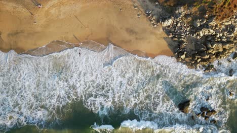 Waves-wash-upon-a-beach-with-people-on-the-beach-below-as-seen-from-a-overhead-aerial-drone