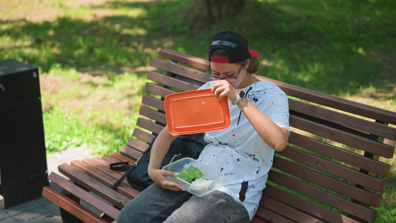 벤치에 앉아 가방에서 샐러드를 꺼내는 통근자, 근처 검은색 쓰레기통, 빨간색 챙 모자, 흰색 얼룩무늬 셔츠, 주황색 용기 열림, 채소 혼합, 음미하는 한 입, 햇빛과 그늘, 편안한 일상, 야외
