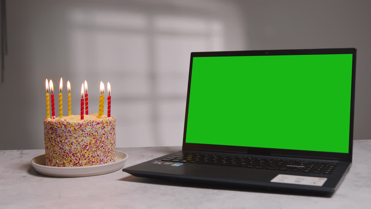 foto de estudio de un portátil de pantalla verde junto a un pastel de cumpleaños cubierto de velas encendidas