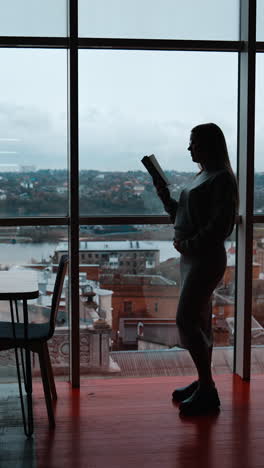 pregnant woman stands near big window focused on book in her hand. long-haired lady reads caressing her belly. cityscape at backdrop. vertical video
