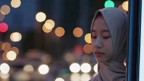 young woman in a hijab standing by a window, gazing thoughtfully as city lights create a serene bokeh backdrop, blending tranquility with urban chaos