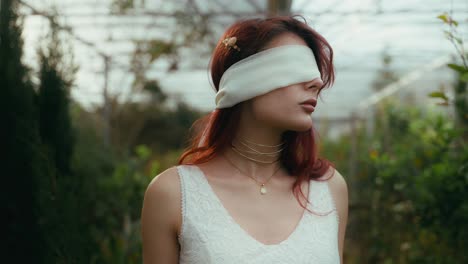 cinematic-circle-pan-footage-of-a-young-woman-in-a-white-dress-and-blindfolded-looking-around-in-a-greenhouse-plant