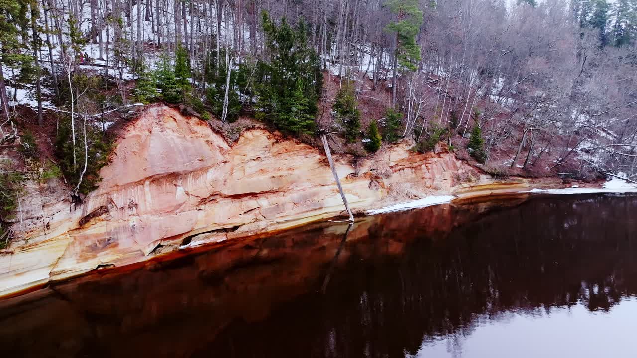 Eroded cliffs of Devil’s Cave glow over Gauja’s dark waters on chilly winter day