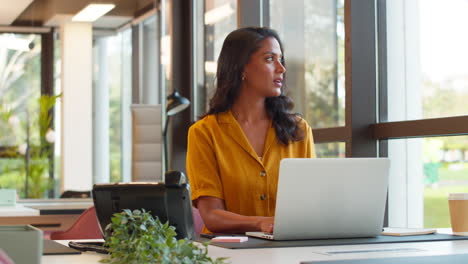 Mature-Businesswoman-Working-On-Laptop-At-Desk-In-Office-Pausing-To-Look-Out-Of-Window