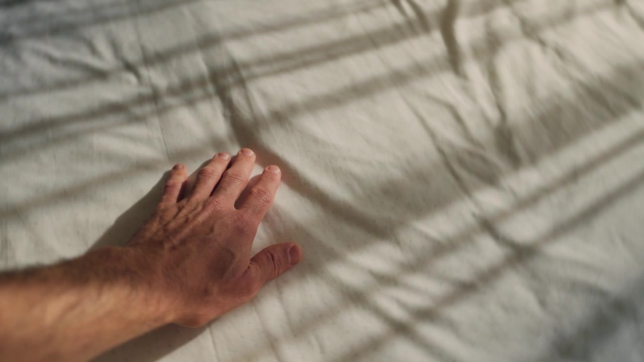 Hand gently caresses bedsheet as sunlight shines into room through window curtains. Comfortable bed. Conceptual abstract shot. Texture of lines and shadows. Romance or memory as hand passes on linen