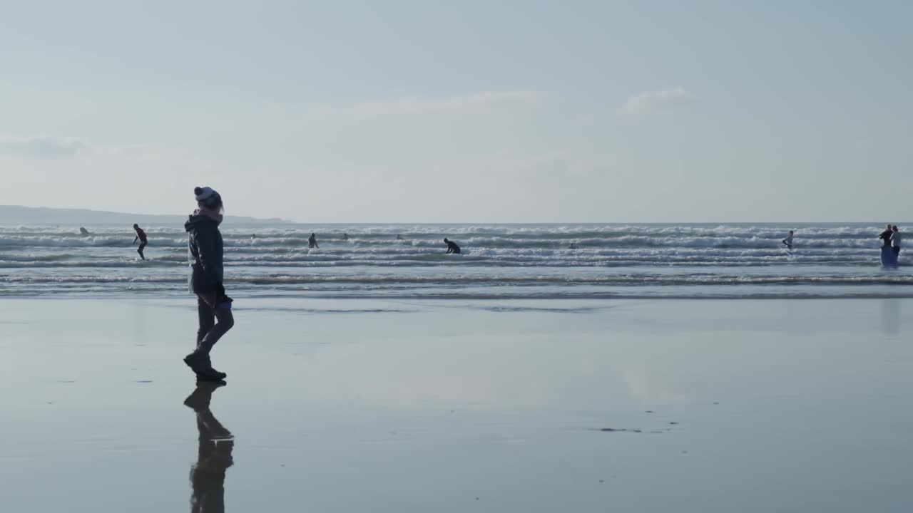 이 완벽한 슬로우 모션 장면에서 서퍼들이 물에 타고 서 있는 동안 워커가 산책하고 있습니다.