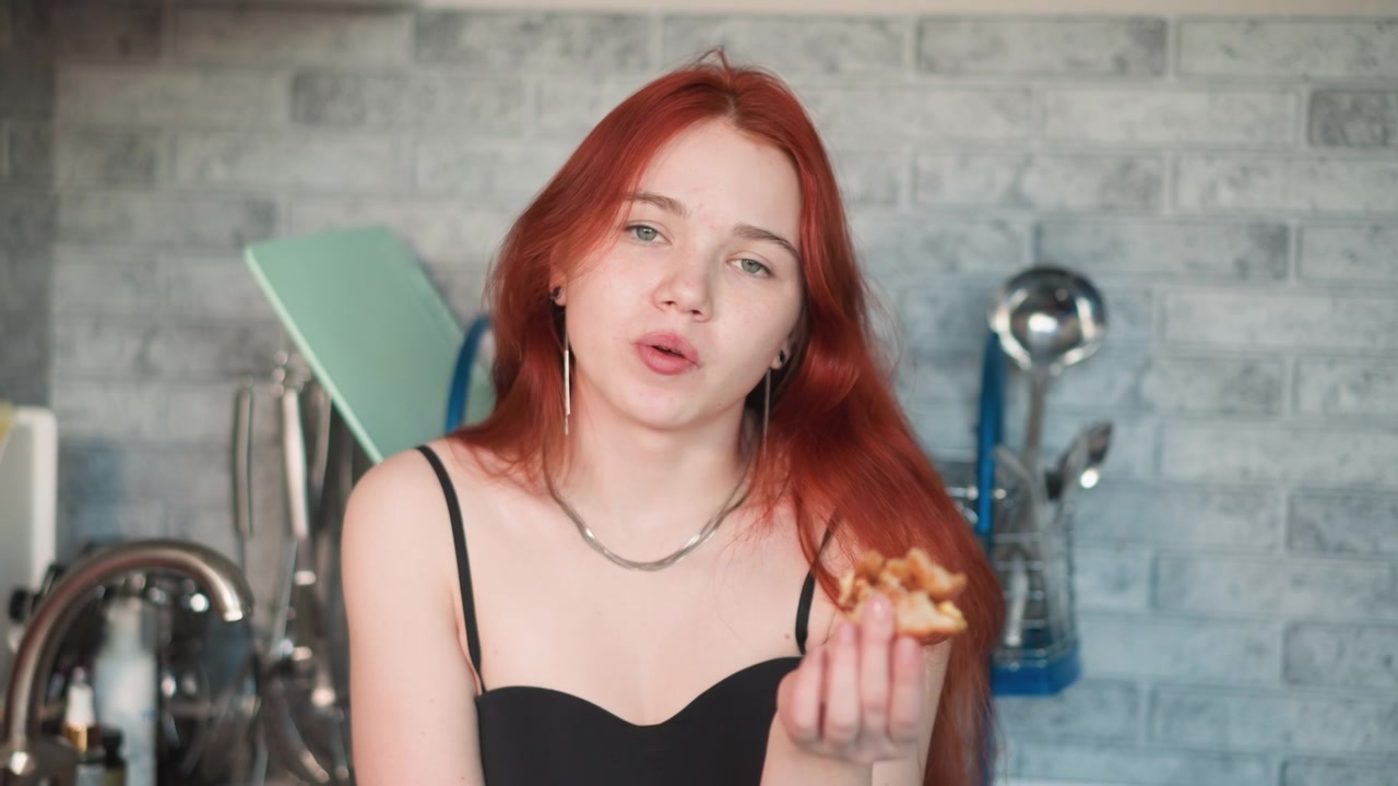 Mujer pelirroja comiendo un pastel, escena de cocina acogedora, masticación juguetona con migas en los dedos, rostro expresivo, compartiendo un bocado con la cámara, luz natural cálida, ambiente de repostería casera, textura mantecosa.