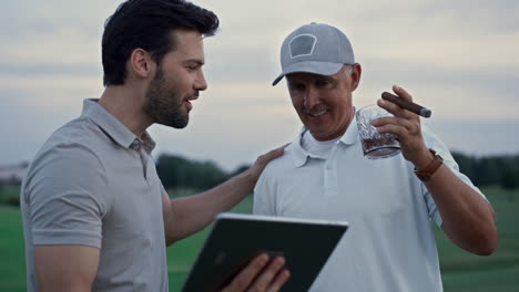 Smiling-men-calling-online-video-holding-tablet-on-golf-course.-Wealthy-concept.