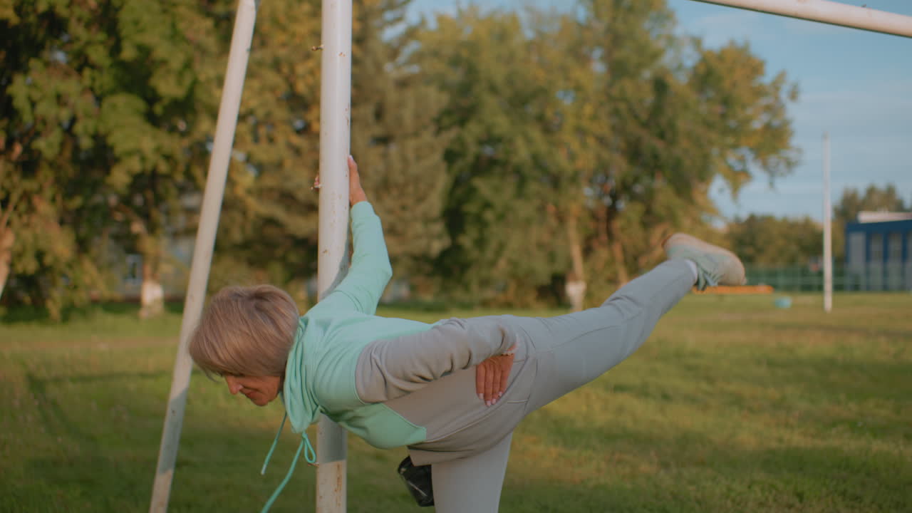 Sports woman holding post performing knee to chest stretch during sunset while puppy tied nearby on grassy field with trees and building in background showcasing outdoor fitness