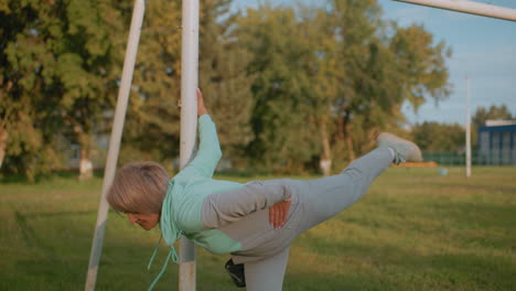sports woman holding post performing knee to chest stretch during sunset while puppy tied nearby on grassy field with trees and building in background showcasing outdoor fitness