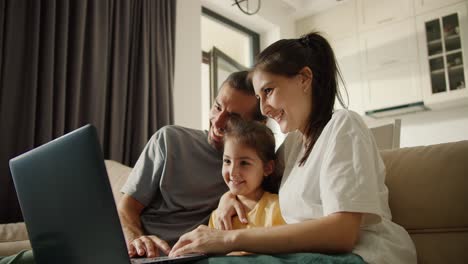 Happy-family,-a-man-with-long-brunette-hair,-his-brunette-girlfriend-and-their-happy-daughter-in-a-yellow-dress-are-sitting-on-a-light-brown-something-in-a-dark-gray-monitor-and-laughing-in-a-modern-studio-room