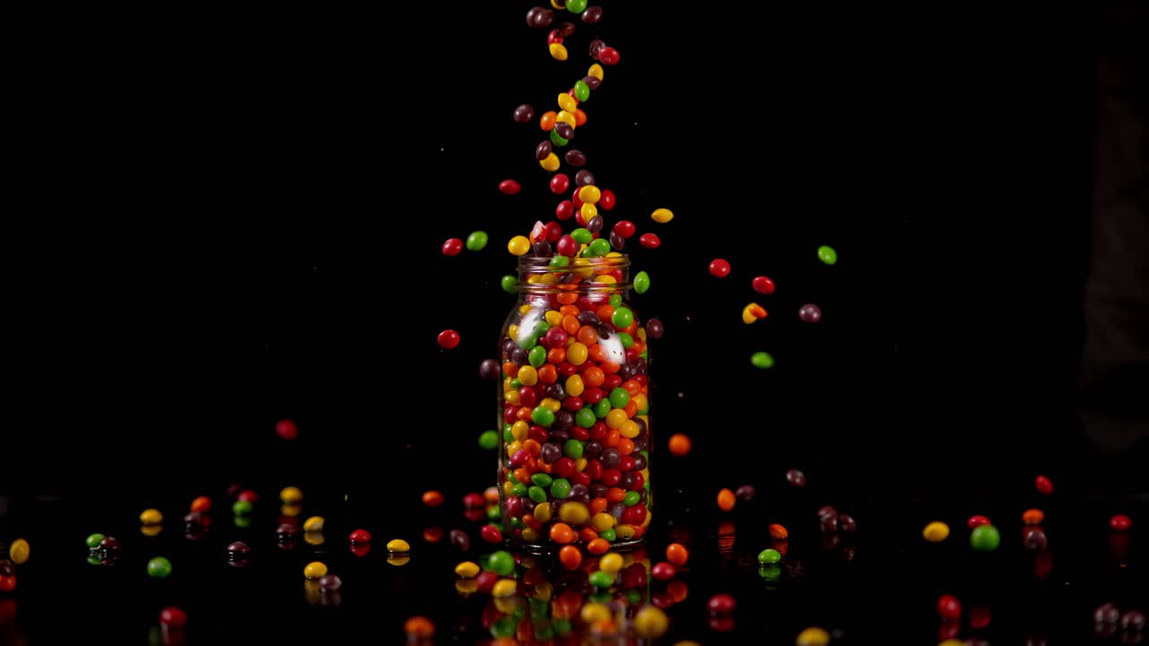 los caramelos de cáscara dura de colores se dejan caer en un frasco de vidrio desbordante en una superficie brillante negra