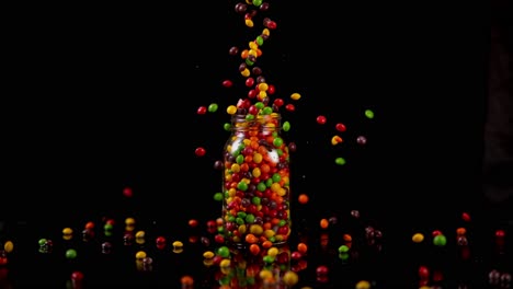 los caramelos de cáscara dura de colores se dejan caer en un frasco de vidrio desbordante en una superficie brillante negra